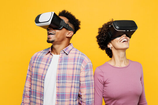 Young fun couple two friends family man woman of African American ethnicity wear purple casual clothes together watching in vr headset pc gadget look aside isolated on plain yellow orange background.