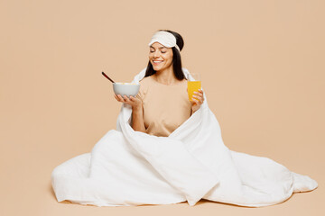Full body young Latin woman wearing pyjamas jam sleep eye mask rest relax at home eat breakfast...