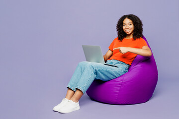 Full body kid teen girl of African American ethnicity wear orange t-shirt sit in bag chair work...