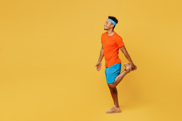 Fototapeta na wymiar Full body young fitness trainer sporty man sportsman wears orange t-shirt raise up leg doing stretch exercise spend time in home gym isolated on plain yellow background. Workout sport fit abs concept.