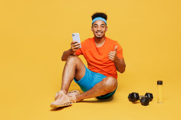 Full body young fitness trainer sporty man sportsman wear orange t-shirt sit hold mobile cell phone show thumb up train in home gym isolated on plain yellow background. Workout sport fit abs concept.