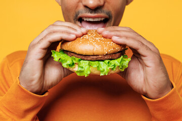 Close up cropped young man wear orange sweatshirt casual clothes holding eating biting burger isolated on plain yellow background studio. Proper nutrition healthy fast food unhealthy choice concept.