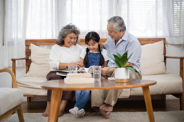 Asian senior couple watching granddaughter reading a book on sofa. Little girl visit her grandparents on holidays weekend. Love and bonding. Healthy family insurance