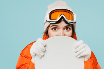 Close up young happy woman wear warm padded windbreaker jacket hat ski goggles mask hold snowboard look camera travel rest spend weekend winter season in mountains isolated on plain blue background.