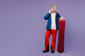 Full body man wear blue windbreaker jacket ski goggles mask hat hold talk speak on mobile cell phone snowboarding spend extreme weekend winter season in mountains isolated on plain purple background.