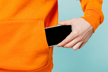Close up cropped young man he wears orange casual clothes hold in hand put into pocket mobile cell phone with blank screen isolated on plain blue cyan color wall background studio. Lifestyle concept.
