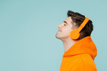 Close up side profile view young man he wearing orange hoody casual clothes listen to music in headphones isolated on plain pastel light blue cyan color background studio portrait. Lifestyle concept.