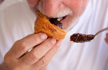 Close up shot of a mature Caucasian senior man biting into a fresh French croissant with cereals...