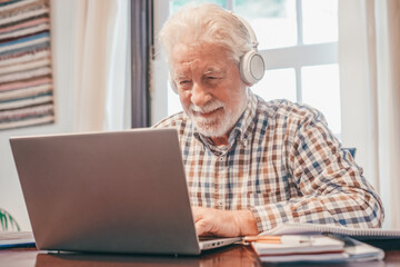 Smiling senior bearded man wearing headphones sitting at table with laptop following online course. Elderly man in checkered shirt enjoying teaching activity and learning using new technology