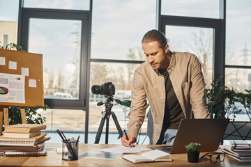 businessman in casual attire writing notes near digital camera and laptop on work desk in office