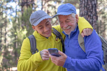 Young grandson and old grandfather consulting trail map on mobile phone while hiking together in mountain forest enjoying nature and healthy activity. Adventure has no age - obrazy, fototapety, plakaty