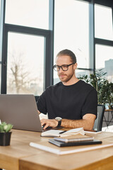 Fototapeta na wymiar stylish man in black t-shirt and eyeglasses typing on laptop at work desk in modern office, business
