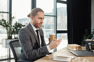 Fototapeta premium serious businessman with smartphone and coffee to go near laptop and notebooks on work desk