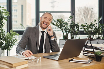 excited successful businessman talking on smartphone and laughing near laptop in modern office