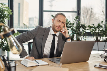 serious stylish businessman sitting at workplace near laptop and looking at camera in office