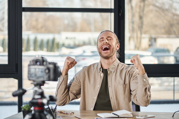 overjoyed businessman showing win gesture in front of digital camera during video blog in office