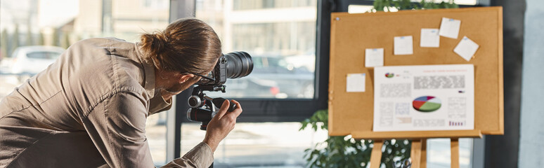 office manager in casual clothes adjusting digital camera near corkboard with infographics, banner