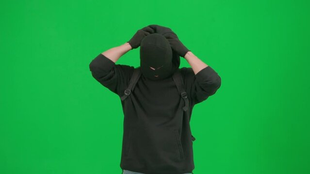 Portrait of thief on chroma key green screen background. Man robber wearing balaclava, checking his gun, fixes backpack getting ready for making a crime.