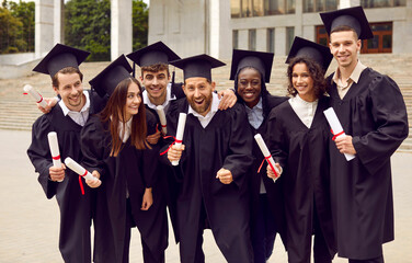 Group of happy excited diverse multiracial multiethnic college or university friends in black student caps and gowns, with diplomas in hands, celebrating their graduation day and having fun together