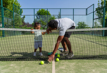 Father teaching his daughter to pick up paddle tennis balls on a tennis court.