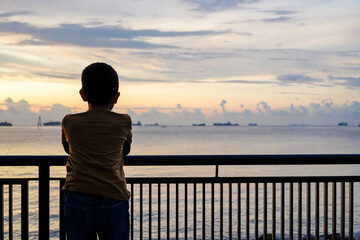 Silhouette of a boy enjoying the view of the sunset and a row of sailing cargo ships