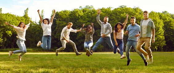 Group of cheerful active friends are jumping up together taking pictures in park in summer. Young multiracial men and women in casual clothes standing in row jumping against background of green trees. - Powered by Adobe