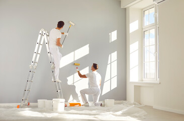 Two male workers from the professional home renovation service painting walls light gray in a big bright living room interior inside a new modern house or apartment