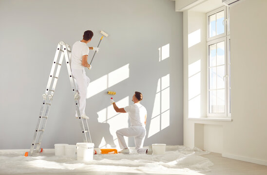 Two male workers from the professional home renovation service painting walls light gray in a big bright living room interior inside a new modern house or apartment