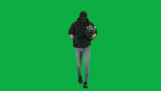 Portrait of thief on chroma key green screen background. Man robber wearing hoodie, jeans and balaclava, running from police with stolen bag full of money.