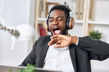 An African-American man sits at his desk in front of his laptop, wearing headphones and chatting on...