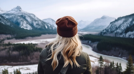 Rearview photography of a carefree young woman looking at the beautiful valleys, snowy mountains, and rivers landscape, outdoors in cold winter, girl with blonde hair wearing a jacket and a cap - obrazy, fototapety, plakaty