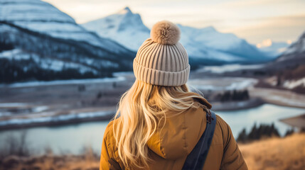 Young woman with blonde hair standing on an Icelandic dry grass field in winter, looking at the snowy mountains and lake or river landscape, wearing a yellow jacket, outdoors winter season, rearview