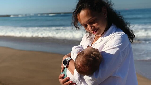 Beautiful curly brunette, loving caring mother holding her baby boy, rocking and kissing him while he's falling asleep in her arms, walking along the beach together on a beautiful warm sunny day