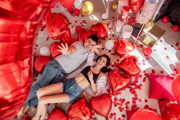 Top view of laughing couple lying on floor, playing with red foil balloons in Valentines Day. Young man and woman hugging, looking each other surrounded by romantic decorations, rose petals. Holiday