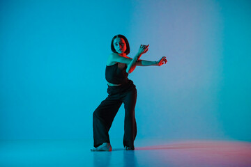 Young woman in black pants and top poses in studio against neon blue background. The dancer...