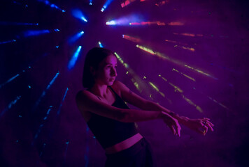 Fototapeta na wymiar A young woman in black casual clothes poses in a studio with smoke, purple light and beams of multicolored light. The dancer demonstrates elements of experimental hip hop style dance choreography.