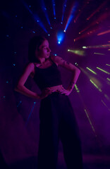 A young woman in black casual clothes poses in a studio with smoke, purple light and beams of...