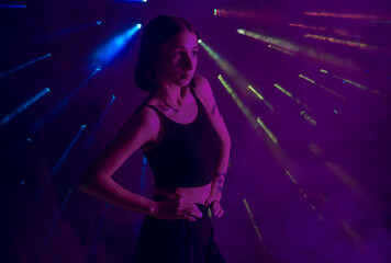 Fototapeta na wymiar A young woman in black casual clothes poses in a studio with smoke, purple light and beams of multicolored light. The dancer demonstrates elements of experimental hip hop style dance choreography.