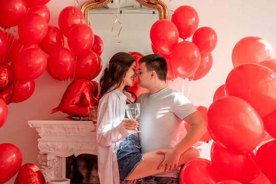 Joyous moment between dancing young couple celebrating with toast Valentines day near red balloons. Woman holding wine glass, covered eyes, man hugs, holds her leg in dance, touch each others noses