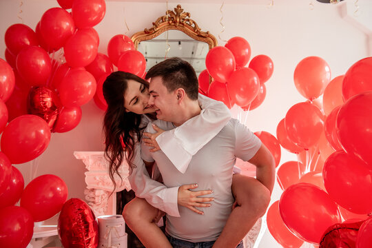 Couple fooling around, laughing, looking into each others eyes. Woman sitting on mans back. Lifestyle holiday Valentines day. Playful, romantic moment near red balloons with white interiors