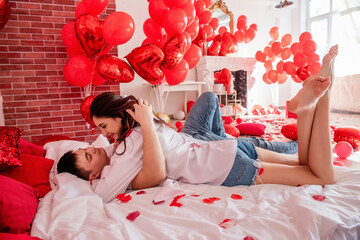 Couple in love hugs tightly on white bed among red balloons, pillows, hearts, rose petals. Man and woman look at each other, spend Valentines Day home in relaxed atmosphere. Surprise gift. Lifestyle