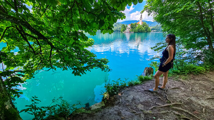 Tourist woman with panoramic view of St Mary Church build on small island on alpine lake Bled, Upper Carniola, Slovenia. Serene landscape in Julian Alps in winter. Hills covered with lush green forest