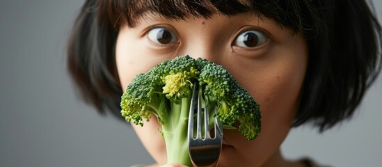 Asian women reluctantly observe broccoli on a fork during mealtime, expressing dislike for vegetables. - obrazy, fototapety, plakaty