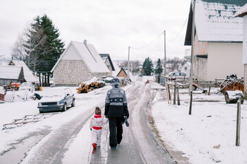 Mom and a little girl walk holding hands along an icy road in a village. Back view
