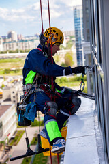 Industrial mountaineering man worker washing glazing, hanging from residential building. Male rope access laborer in work uniform during high-rise job. Industry urban works concept. Copy ad text space
