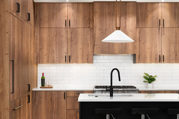A kitchen detail with wood cabinets, a black faucet, subway tile backsplash, and gold light fixtures hanging above the black island and marble countertops. - obrazy, fototapety, plakaty