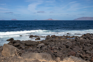 Detail of a textured dark brown to black volcanic stones on the coast of Atlantic ocean. Mountain in the background. Blue sky with some white clouds. Central Lanzarote, Canary Islands, Spain.