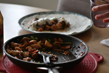 Close up of a busy dining table focused on a pan of fajita chicken. A plate with a chicken wrap is scene in the background with a sauce bottle and a glass on the sides.