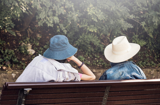 Rear view with two women wearing sun hats sitting on a bench in the park.