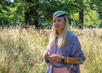 A sweet young woman with blond hair, wearing a scarf on her head, stands in the tall grass, looking at the camera. People outdoors © Alexander